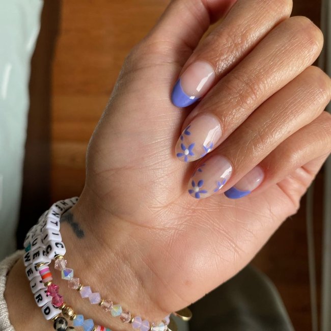 Nail art com francesinha roxa Mão com os dedos dobrados e pulseiras coloridas no pulso, exibindo as unhas com francesinha roxa e flores roxas.