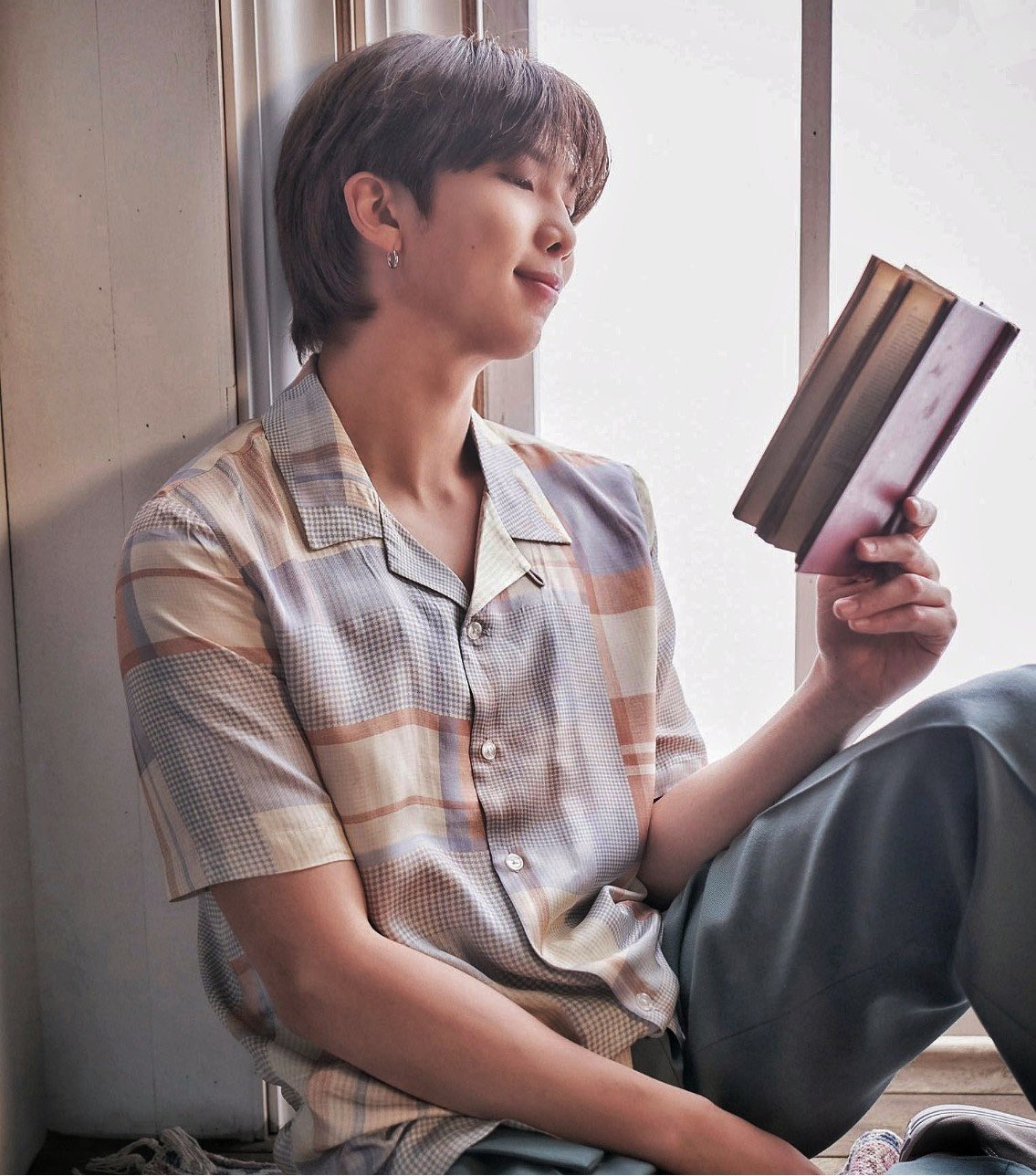 RM do BTS sentado em uma janela lendo um livro, ele segura o livro com apenas uma das mãos e a outra está apoiada na perna