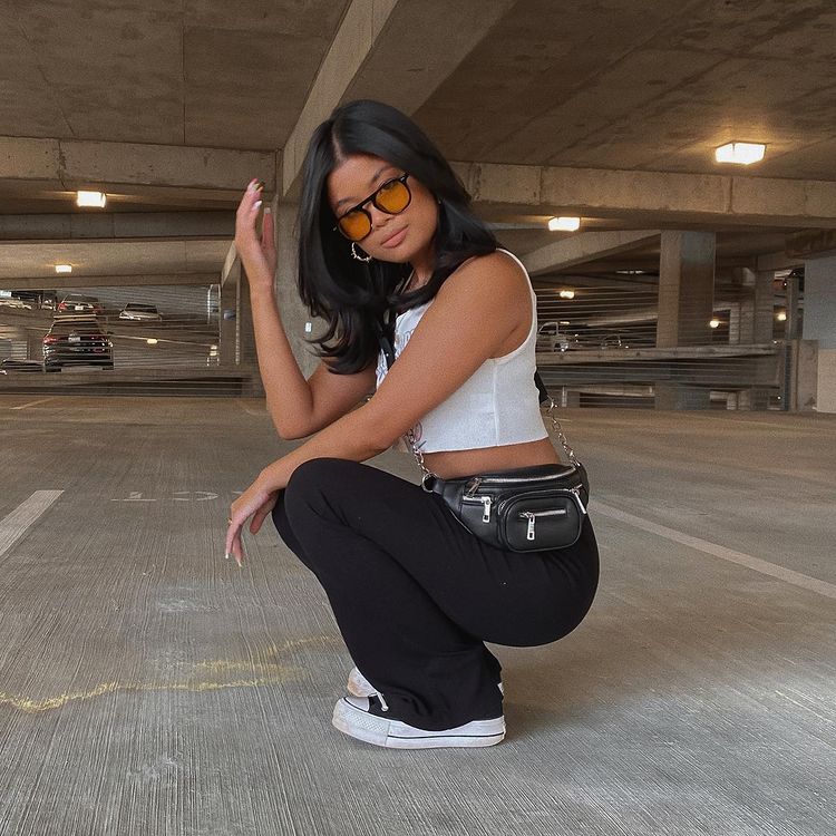 aly_calca_bailarina Foto de uma mulher em um estacionamento. Ela usa um cropped branco, calça bailarina preta, pochete e óculos de sol com lente amarela. Ela está agachada, olha para a câmera e não sorri.