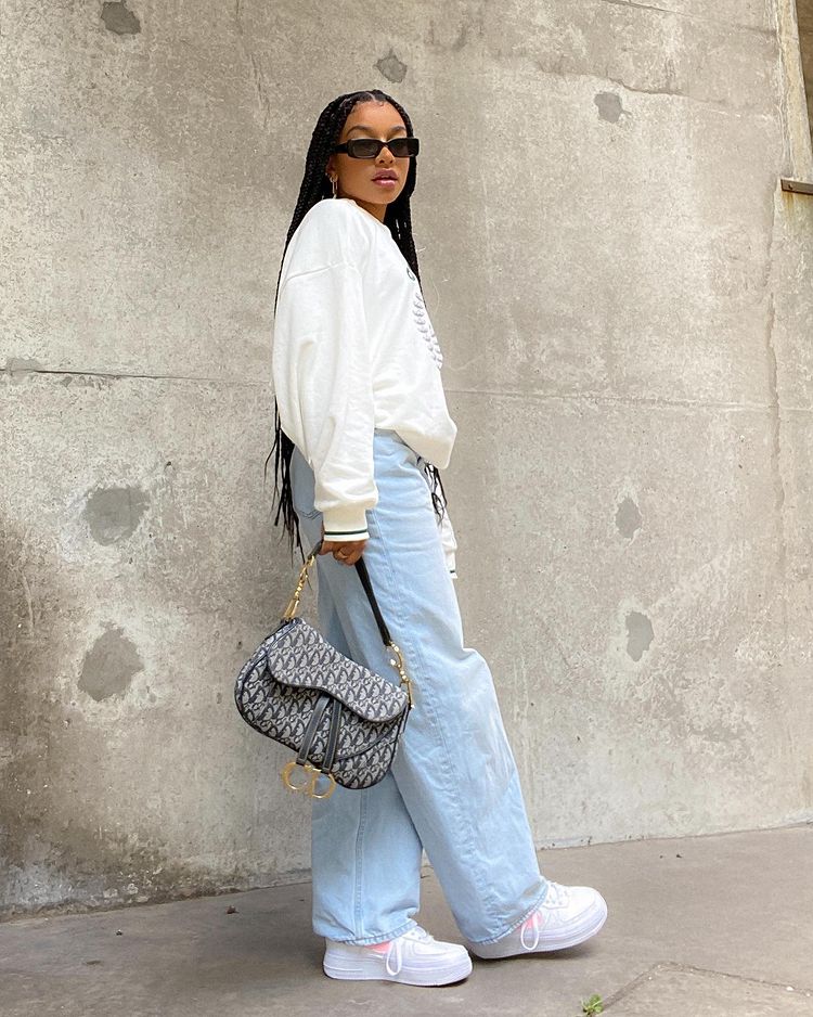 Foto de uma mulher em uma calçada. Ela usa um moletom branco, calça jeans baggy, tênis branca, shoulder bag e um óculos de sol vintage. Ela olha para a câmera e não sorri.