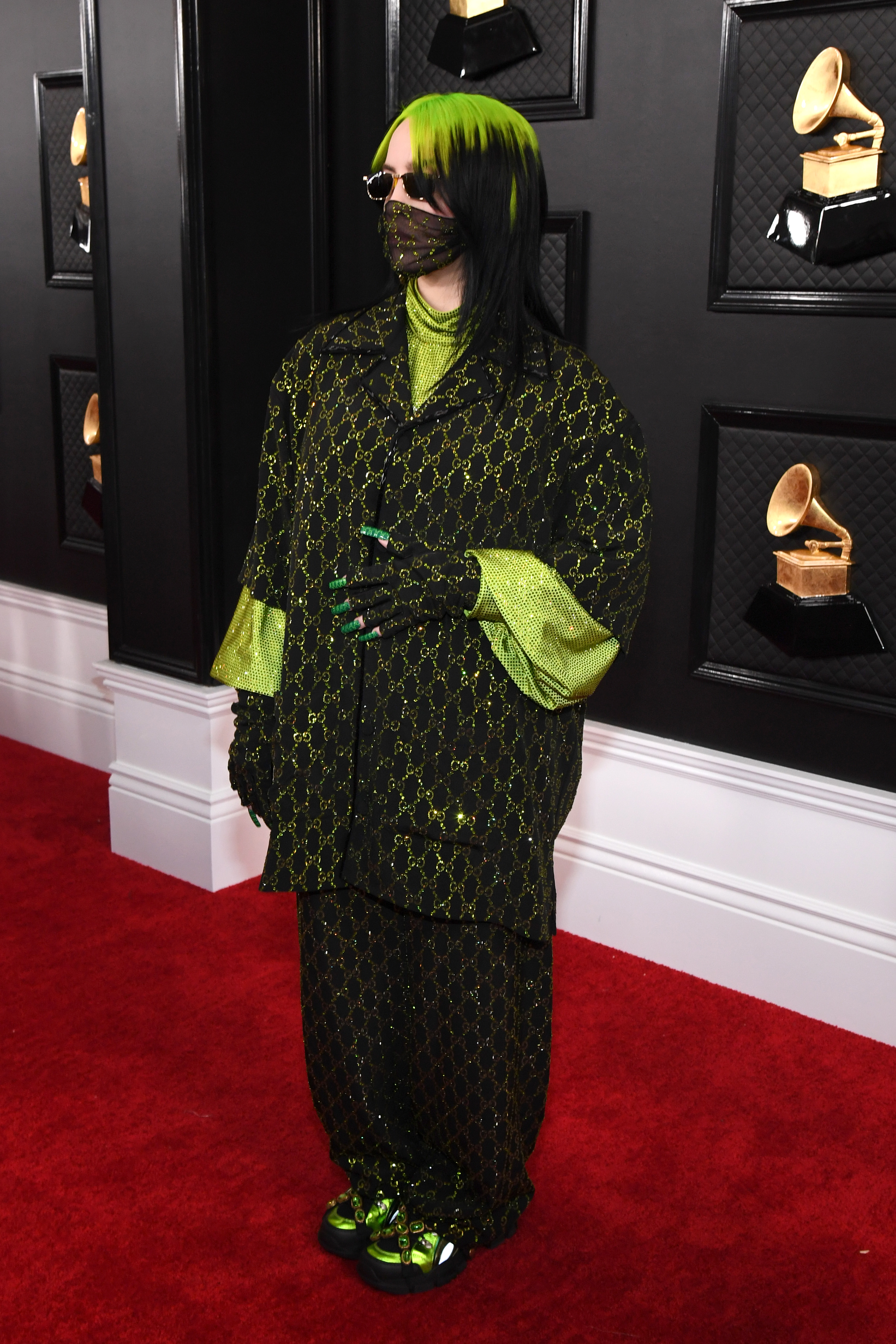 Foto da cantora Billie Eilish no tapete vermelho do Grammy Awards. Ela usa uma camisa oversized preta com detalhes verdes, calça preta com detalhes verdes e tênis preto com detalhes verdes. Ela não olha para a câmera.