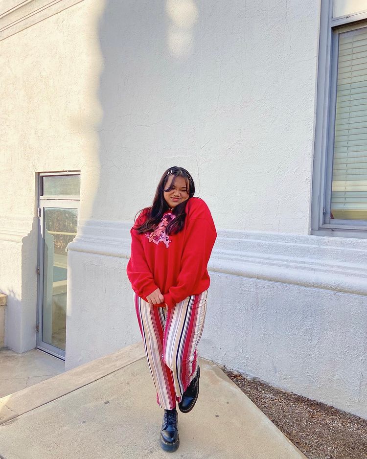 Foto de uma mulher em frente a uma casa de muro branco. Ela usa um moletom vermelho baggy, calça com listras brancas, vermelhas, pretas e bege verticais e coturno. Ela está de olho fechado e sorri para a foto.
