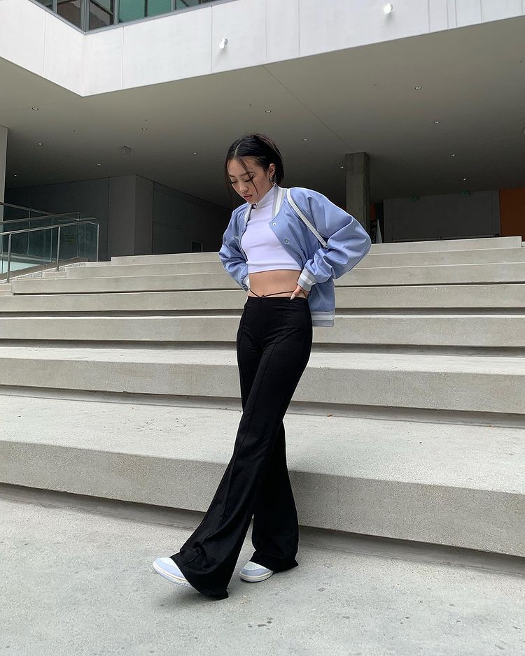 calca_legging_bailarina Foto de uma mulher em uma calçada. Ela usa um top cropped branco de gola alta, jaqueta bomber azul, calça legging bailarina preta e tênis azul com branco. Ela está com as mãos na cintura, o pé esquerdo levemente levantado e olha para o chão.