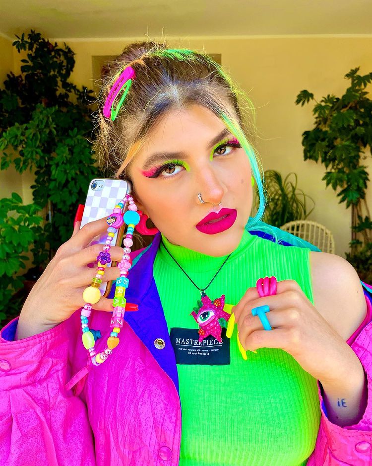 Garota usando top cropped verde neon e jaqueta roxa e azul. Ela está segurando seu celular, que tem uma cordinha de miçangas pendurada, perto da orelha, e sua expressão facial é séria. Sua maquiagem é verde neon e rosa, seu cabelo está preso e há presilhas rosa e verde neon na cabeça.