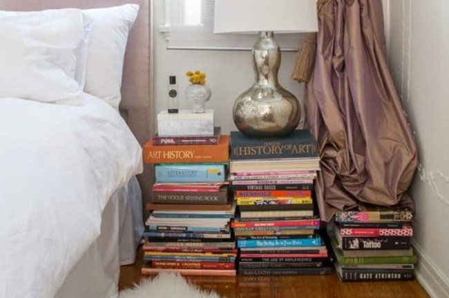 8 invenções que você fez no quarto, mas se arrependeu totalmente depois Quarto branco com uma mesa de cabeceira feita com pilhas de livros coloridos