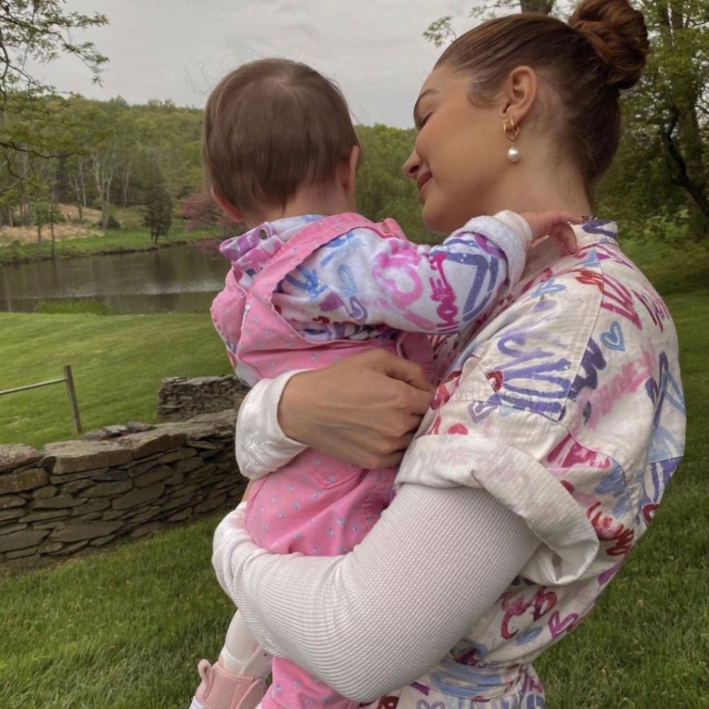 Gigi Hadid segurando a filha Khai no colo em um campo de área verde; as duas vestem roupas combinando em tons de rosa, roxo, branco e azul; Khai está de costas para câmera e Gigi está olhando para ela e sorrindo