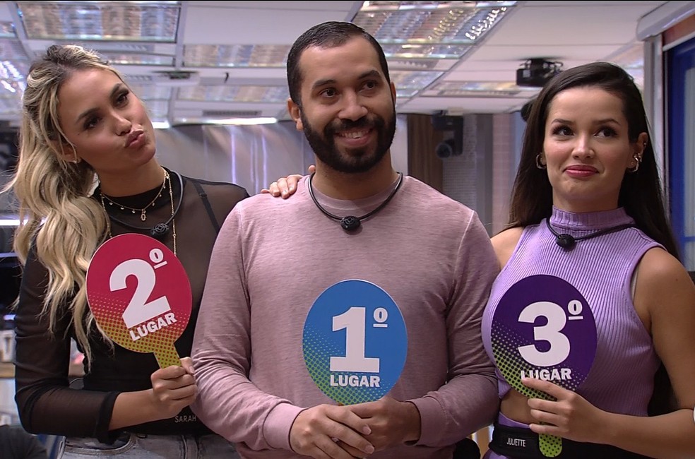 Gil, ao centro, segura a placa de primeiro lugar sorrindo. À esquerda, Sarah segura a placa de segundo lugar. À direita, Juliette segura a placa de terceiro lugar