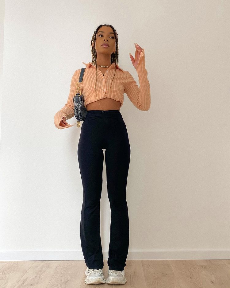lisa_calca_legging_bailarina Foto de uma mulher. Ela usa uma camisa cropped de manga longa, calça bailarina preta, tênis branco e shoulder bag preta. Ela não olha para a câmera e não sorri.