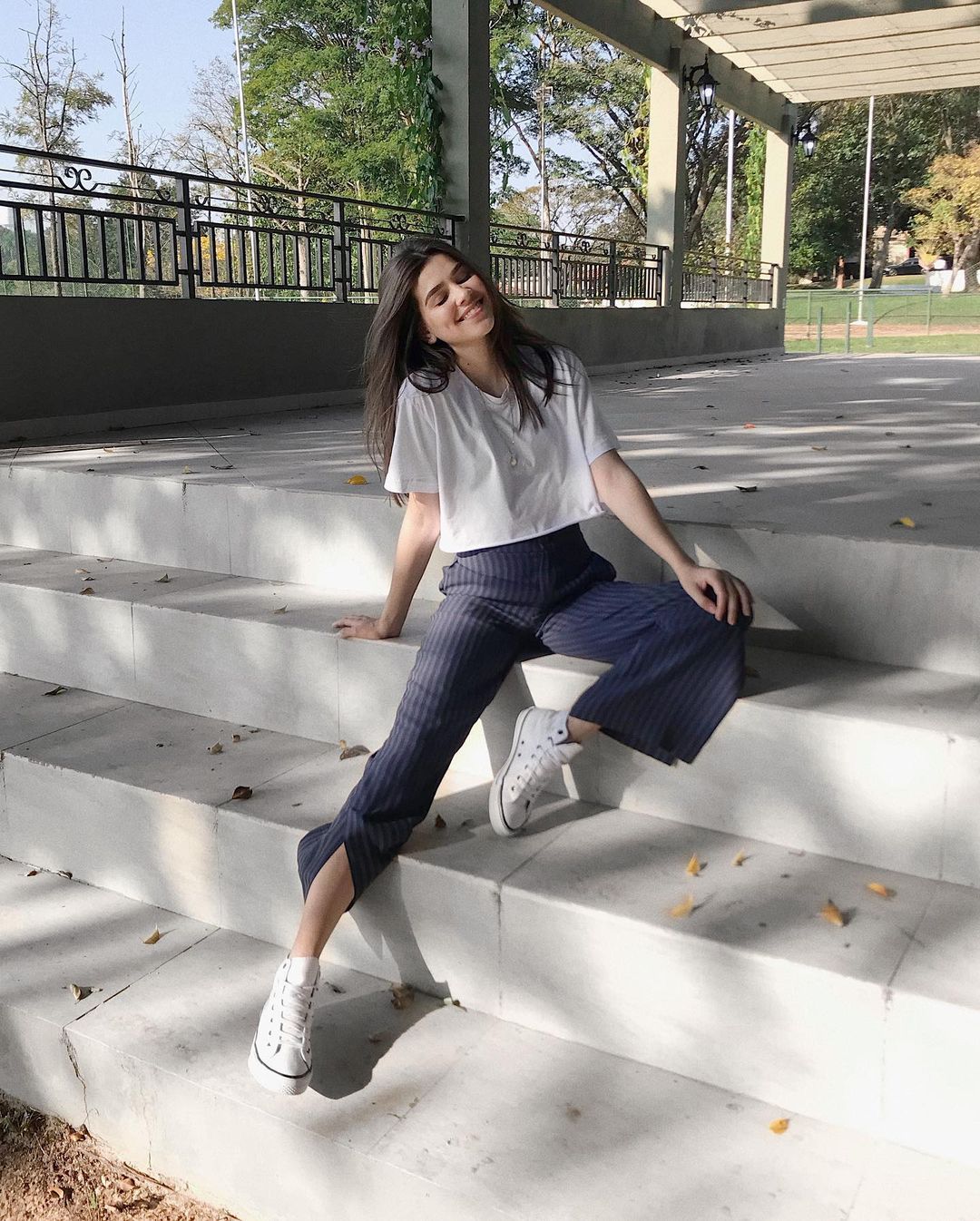 A mulher está sentada em uma escada. Ela veste uma camiseta branca, calça de alfaiataria azul e tênis de cano alto branco. Ela está de olhos fechados sorrindo.