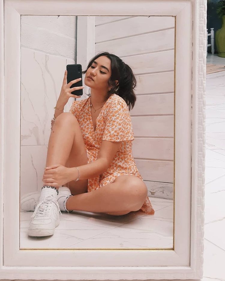 look-vestido-tenis-branco Selfie de uma garota no espelho sentada no chão. Ela usa um vestido laranja com flores brancas, tênis branco e está com o cabelo preso. Ela olha para o celular e não sorri para a foto.