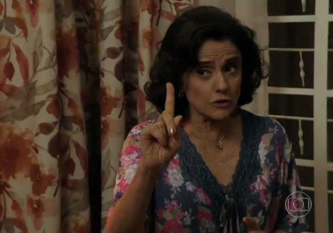 mae Marieta Severo interpretando Dona Nenê. A atriz usa uma blusa estampada e está com o dedo indicador levantado. Ao fundo, aparece uma cortina florida.