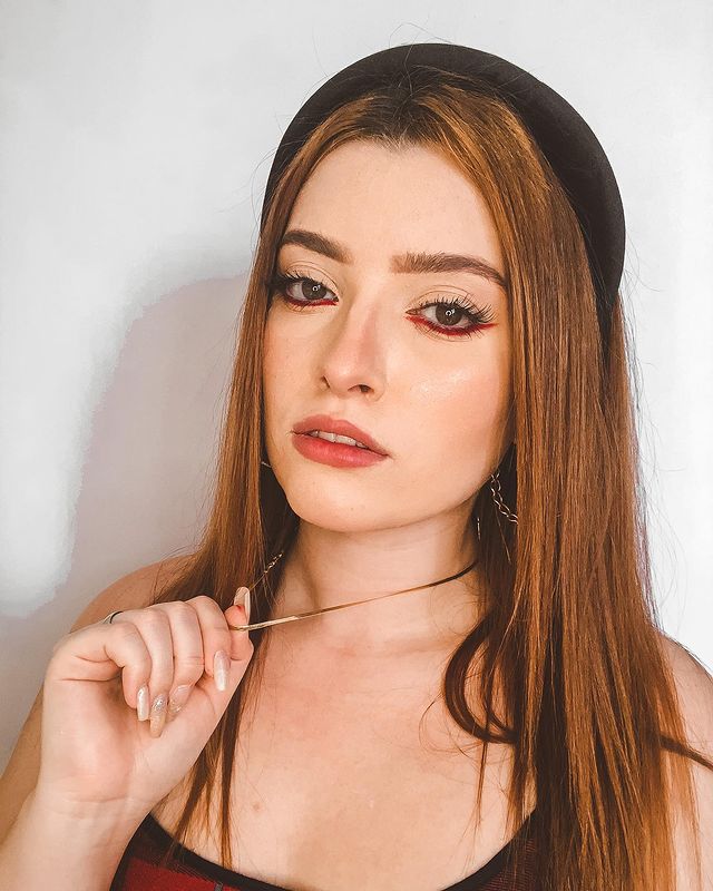 maquiagem-delineado-invertido-limmonada Garota ruiva de cabelo solto usando uma tiara preta de veludo e uma maquiagem com delineado vermelho invertido rente aos cílios inferiores. Ela está com expressão facial séria e uma das mãos puxando seu colar dourado.