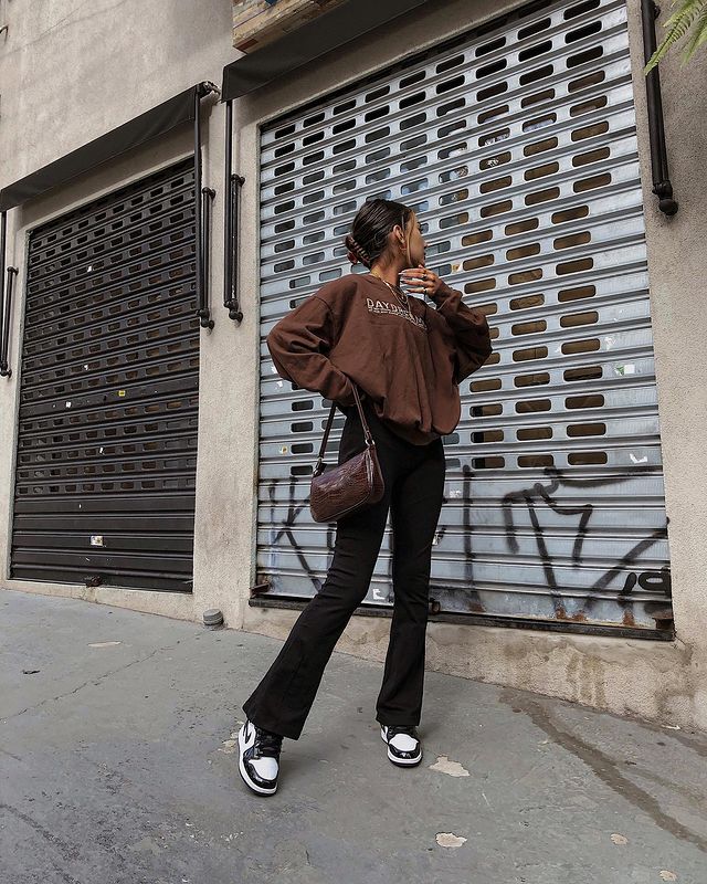 moletom_vintage_com_calça Foto de uma mulher na calçada. Ela veste um moletom vintage marrom, calça bailarina preta e tênis preto e branco. Ela olha para o lado e faz pose.