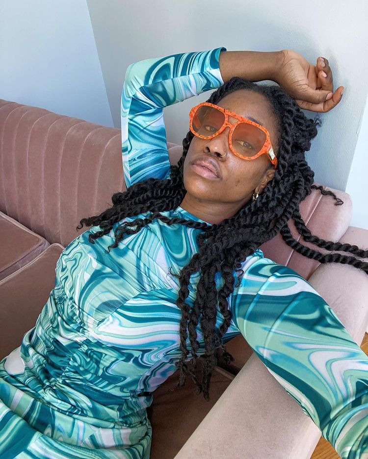 oculos-de-sol-lente-laranja-thelanguageofyolande Mulher sentada em um sofá com a cabeça apoiada e inclinada para cima, com uma das mãos na cabeça. Ela está usando um vestido de manga comprida em tons de azul, um óculos de sol laranja e sua expressão facial é séria.