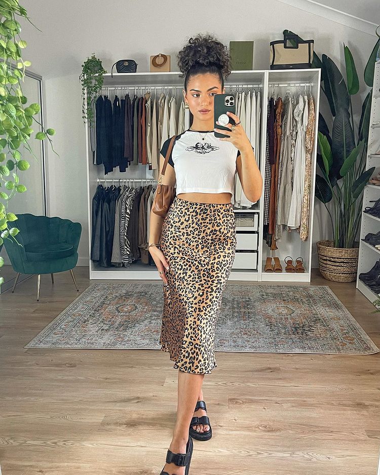 saia-animal-print-oliviacalabio Garota usando camiseta cropped branca e preta com saia midi de estampa de alcinha, sandália preta e uma bolsa pendurada em um dos ombros. Ela está tirando a foto em frente ao espelho com o celular próximo ao rosto e sua expressão facial é séria.
