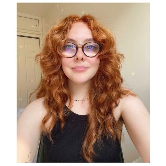 Curtain bangs com cabelo ruivo Jovem sorridente usando óculos e regata preta. Ela posa fazendo uma selfie e atrás dela vemos uma porta branca.