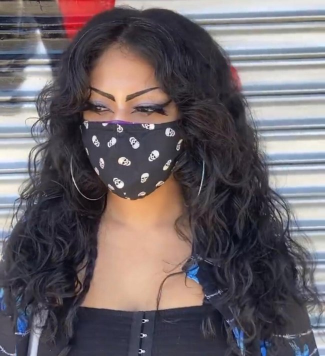 Curtain bangs Jovem olhando para baixo com máscara preta e blusa xadrez azul com camiseta preta por dentro. Sua expressão é séria.