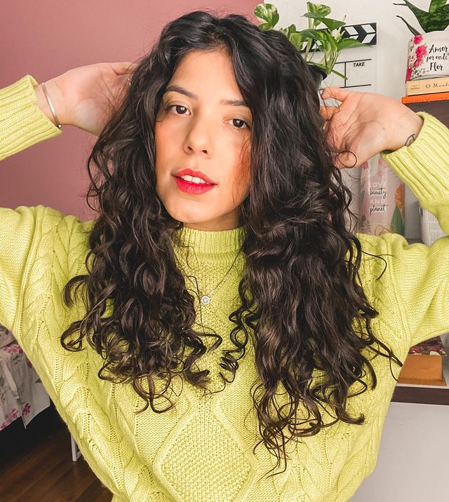 Curtains bangs cabelo cacheado castanho Jovem posa com as duas mãos apoiadas na cabeça, usando blusa amarela esverdeada, sua expressão é séria.