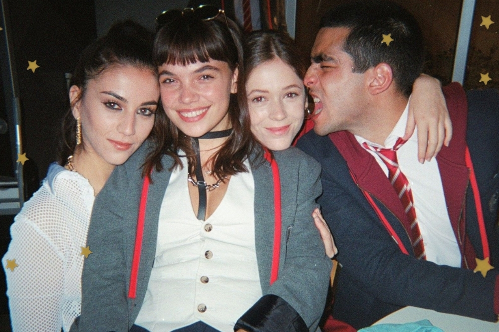 Rebeca, Mencia, Caye e Omar do seriado Elite, sentados com o uniforme da escola e sorridentes, posando para a foto. Omar está com o rosto virado para Caye.