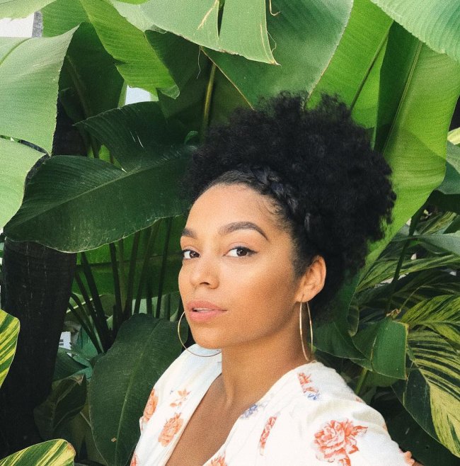 Jovem posando em frente a folhagens verdes, com expressão séria e usando blusa branca com estampas florais.