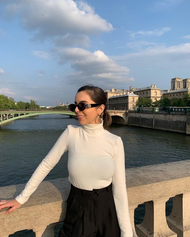 blusa branca de gola alta Foto de uma mulher em frente a um rio em Paris. Ela usa uma calça preta de alfaiataria, blusa branca de gola alta e manga longa. Olha para o lado e sorri.