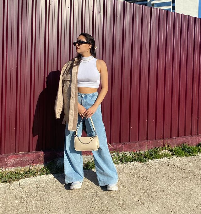 blusa de gola alta branca Foto de uma mulher na calçada. Ela usa uma blusa branca de gola alta sem manga, calça wide leg, tênis branco, casaco de veludo bege e bolsa bege. Ela olha para o lado e não sorri.