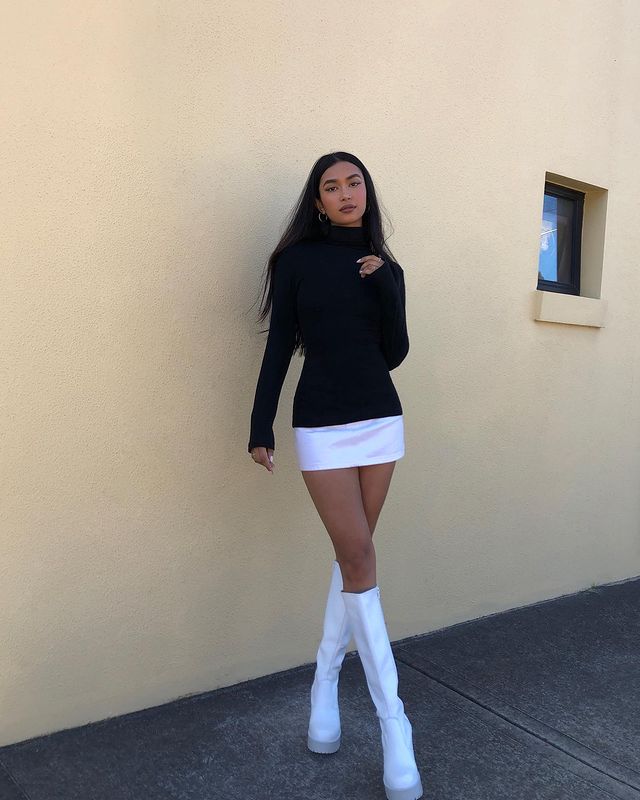 blusa de gola alta preta Foto de uma mulher na frente de uma parede rosa. Ela usa uma blusa preta de gola alta e manga longa, saia branca e bota de cano alto branca. Ela olha para a câmera e não sorri.