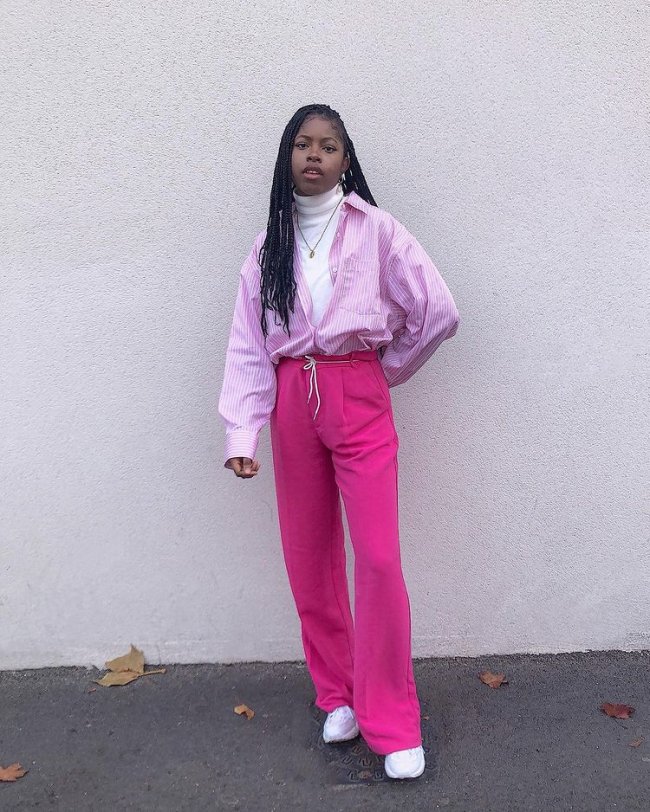 blusa de gola alta Foto de uma mulher na rua. Ela usa uma blusa de gola alta branca, camisa rosa com listras brancas aberta, calça rosa e tênis branco. Ela olha para a câmera e não sorri.