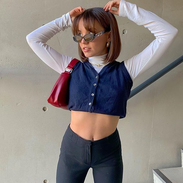 blusa gola alta branca Foto de uma mulher perto de uma escada. Ela usa uma calça preta, blusa branca de gola alta, camisa sem manga como sobreposição, bolsa vermelha e óculos de sol. Ela está com os braços levantados e mão atrás da cabeça, ela olha para o lado e não sorri.