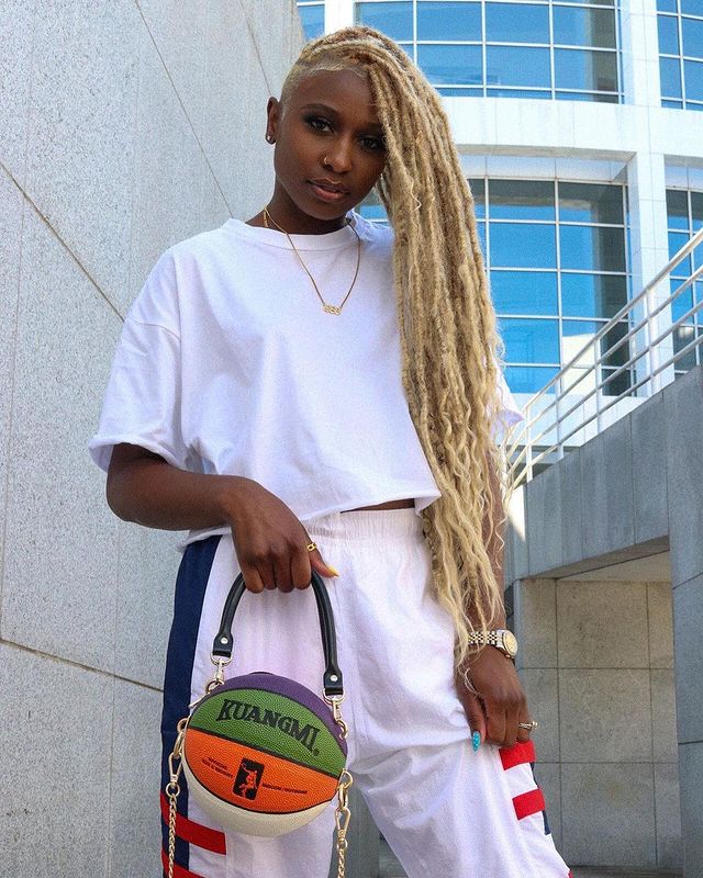 bolsa formato bola de basquete Foto de uma mulher perto das rampas de um edifício. Ela usa uma camiseta branca, calça de moletom rosa clara e segura a bolsa em formato de bola de basquete na mão direita. Ela olha para a câmera e não sorri.