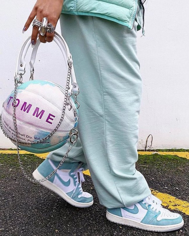 bolsa bola de basquete azul Foto de uma mulher da cintura para baixo. Ela usa uma calça moletom azul, tênis com detalhes brancos e azul e segura uma bolsa branca e azul em formato de bola de basquete.