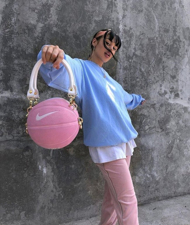 Foto de uma mulher encostada em um muro. Ela usa uma camiseta oversized branca, moletom azul como sobreposição, calça rosa e bolsa em formato de bola de basquete rosa. Ela está de braços abertos, olha para a câmera e não sorri.