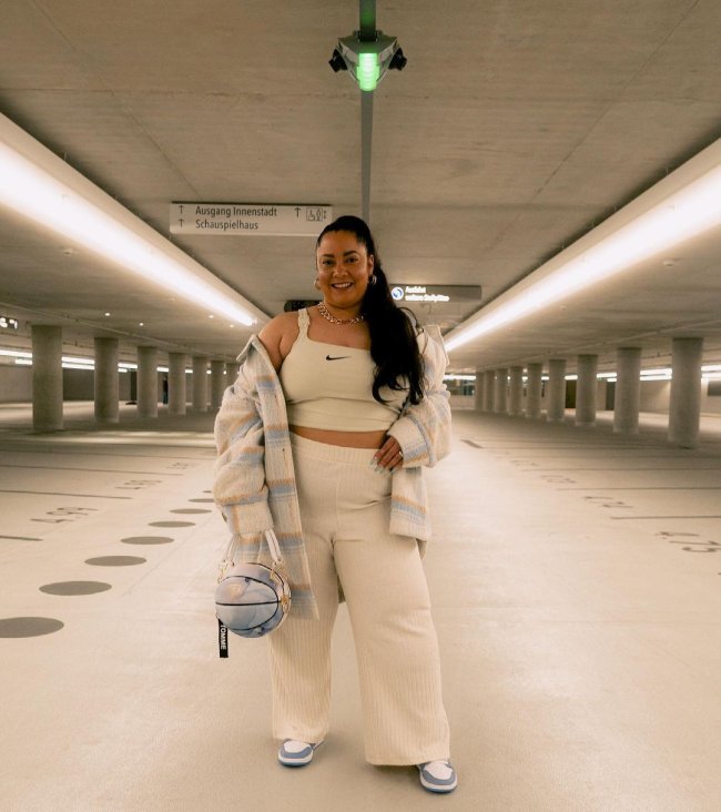 bolsa bola de basquete Foto de uma mulher no estacionamento. Ela usa um top e uma calça na cor creme, tênis nas cores azul e branca e segura a bolsa em formato de bola de basquete na mão direita. Ela olha para a câmera e sorri.