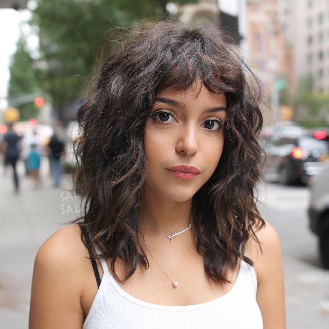 cabelo curto com curtain bangs Jovem com expressão séria posando no meio da rua, podemos ver pessoas e árvores desfocadas atrás dela. Ela usa uma regata, a imagem só mostra dos seus seios para cima.