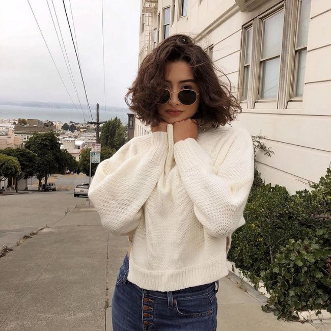 cabelo ondulado corte reto Foto de uma mulher na calçada. Ela usa uma calça jeans, casaco branco, óculos de sol e cabelo ondulado solto em um corte reto acima do ombro. Ela olha para a câmera e não sorri.