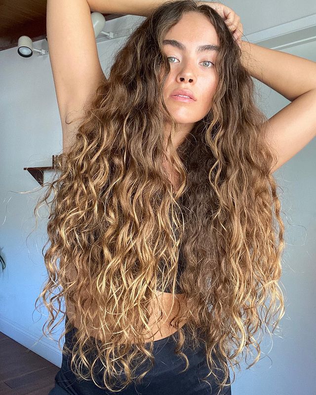 cabelo ondulado corte u Foto de uma mulher. Ela usa uma calça e top preto e o seu cabelo longo e ondulado está solto. Ela está com as mãos na cabeça, olha para a câmera e não sorri.