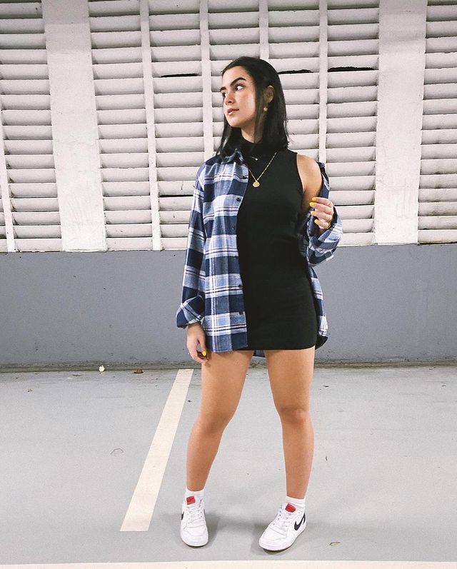 Foto de uma menina em um estacionamento. Ela usa um vestido preto de gola alta e uma camisa xadrez oversized como sobreposição. Ela olha para o lado e não sorri.