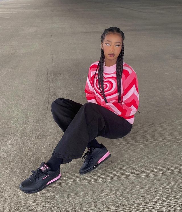 casaco de gola alta Foto de uma mulher sentada no chão de um estacionamento. Ela usa blusa rosa de gola alta com estampa de coração, calça preta e tênis preto. Ela olha para a câmera e não sorri.