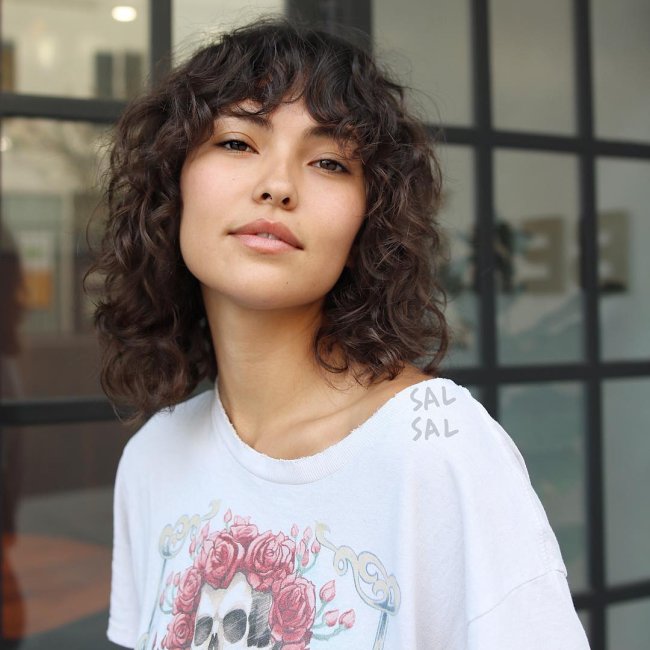 cortininha cabelo cacheado Jovem com a cabeça levemente inclinada, usando camiseta com estampa de coroa. Sua expressão é sorridente.