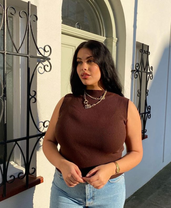 cropped gola alta Foto de uma mulher em uma calçada. Ela usa uma calça jeans, uma blusa marrom sem manga e de gola alta. Ela olha para o lado e não sorri.