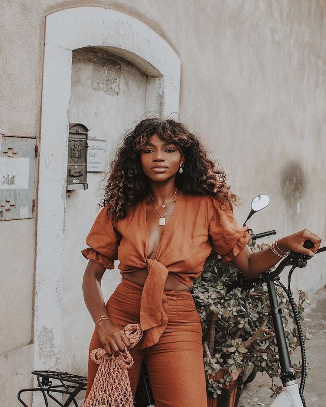 curtain bangs cabelo Jovem posando com look laranja monocromático, em apoiada em uma bicicleta com flores, ainda conseguimos ver uma porta branca atrás dela.
