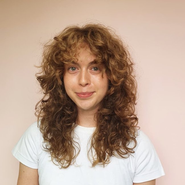 curtain bangs cabelo com cachos Jovem posando sorridente com camiseta branca e em frente a parede bege.