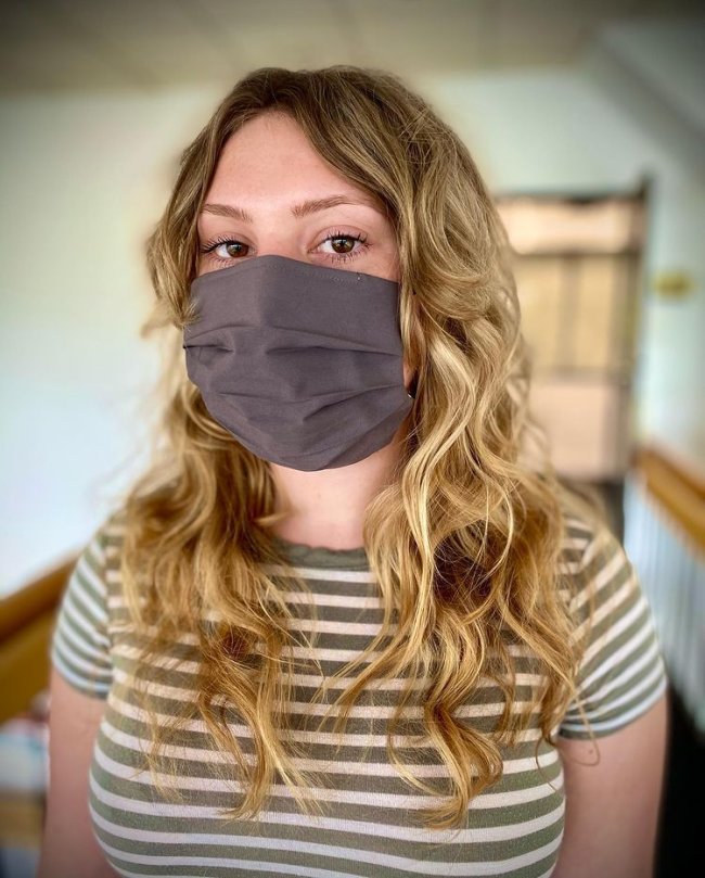 curtain bangs cabelo loiro e cacheado Jovem usando máscara de proteção preta e cobrindo sua expressão facial. Ela veste uma camiseta listrada.