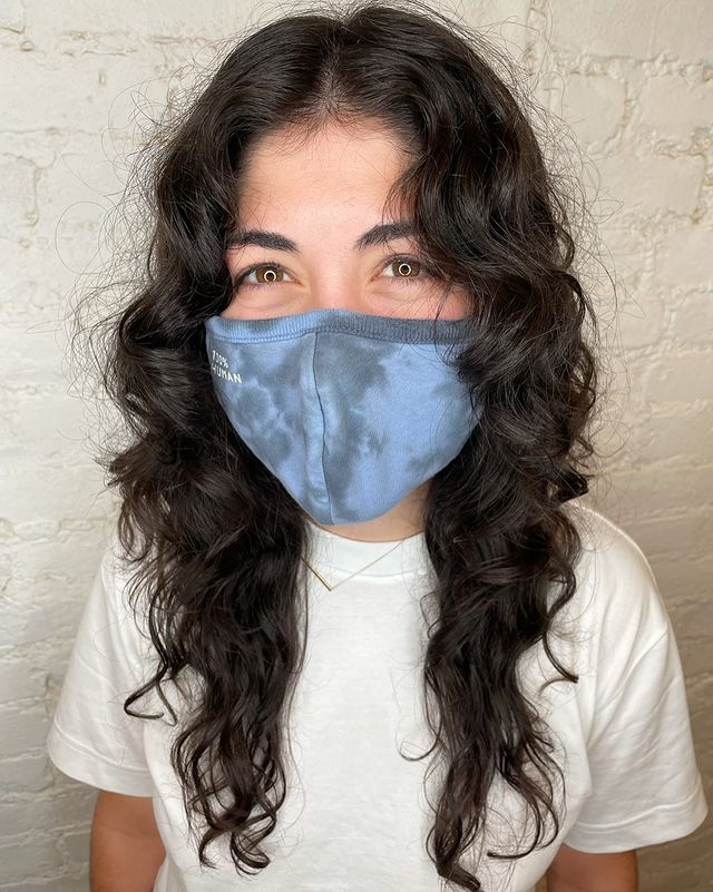 curtain bangs com cabelo castanho cacheado Jovem usando máscara de proteção azul e cobrindo sua expressão facial. Ela veste uma camiseta branca, pelo seu olhar parece sorridente.