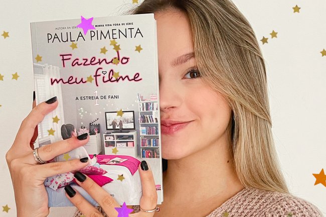 Bela Fernandes segurando o livro Fazendo Meu Filme e escondendo metade do rosto atrás dele; ela está sorrindo levemente com maquiagem clara e as unhas pintadas de preto em um fundo branco/neutro; estrelas amarelas, laranjas e lilás decoram a imagem