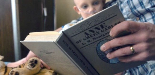 hopper-stranger-things-lendo Ima mão, com uma alianaça, segura um livro. Ao fundo, uma menina, com aparelho respiratório, está sentada no colo da pessoa que lê e olha para o livro
