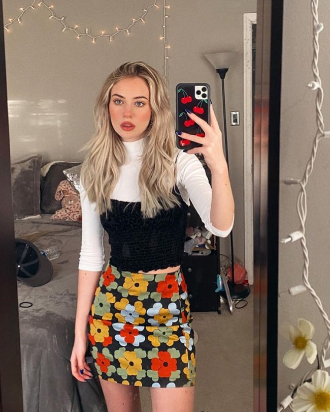 look com blusa branca Selfie no espelho de uma mulher. Ela usa uma saia de flores colorida, blusa branca de gola alta e cropped preto por cima. Ela olha para o espelho e não sorri.