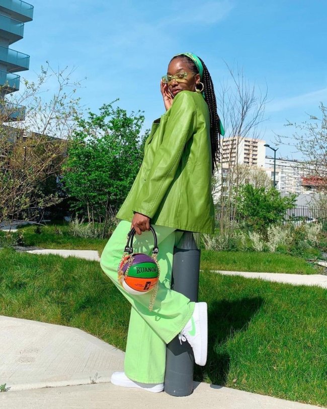 look com bolsa formato bola de basquete Foto de uma mulher em um parque. Ela usa um casaco verde, uma calça verde, tênis branco e bolsa em formato de bola de basquete. Ela está virada de lado, com a perna esquerda levemente levantada, está com a mão direita segurando o óculos verde no rosto, olha para a câmera e não sorri.