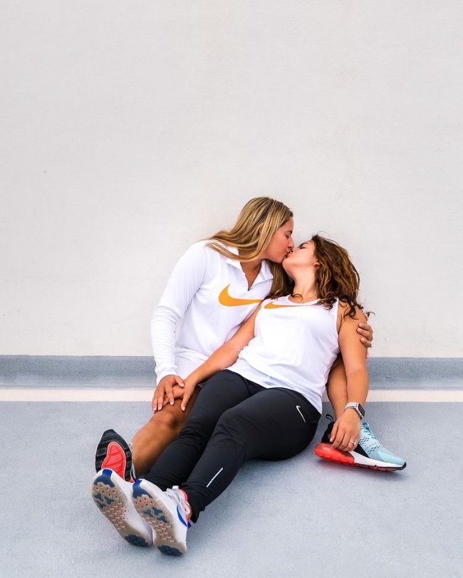 Casal deitado no chão usando roupas para academia da Nike, elas estão se beijando e não conseguimos ver seus rostos. Uma está com uma camiseta branca com manga longa da nike e a outra com regata branca da Nike e legging.