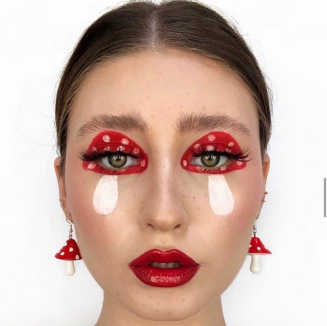 maquiagem de cogumelo Foto do rosto de uma mulher em um fundo branco. Ela está com uma maquiagem em formato de cogumelo nos olhos, batom vermelho, cabelo preso em um coque baixo e brincos de cogumelo. Ela olha para a câmera e não sorri.